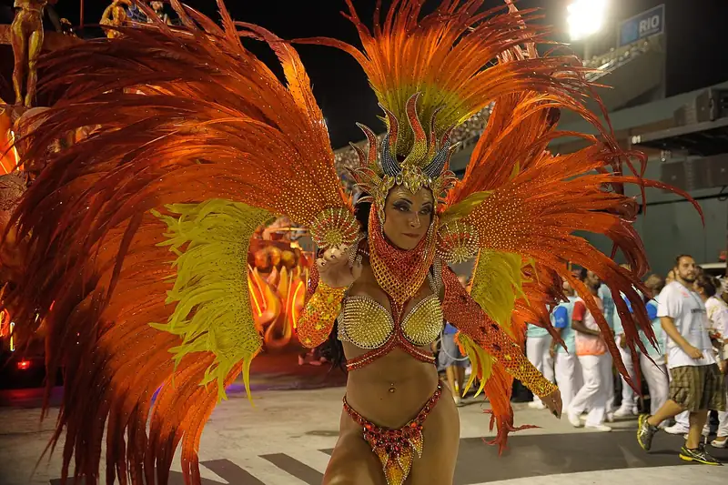 Une danseuse de samba en costume de plumes orange et or défile sur la piste, les tribunes en arrière-plan