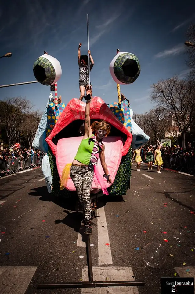 Une acrobate de Cirkul’R pose devant un char en forme de bouche géante aux lèvres rouges et aux gros yeux, pendant qu'une autre danse à la barre au sommet, au milieu d'un défilé