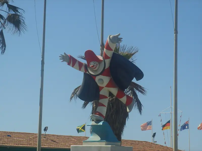 Statue de Burlamacco perchée sur un mât, bras ouverts, en costume à damier rouge et blanc avec une cape noire