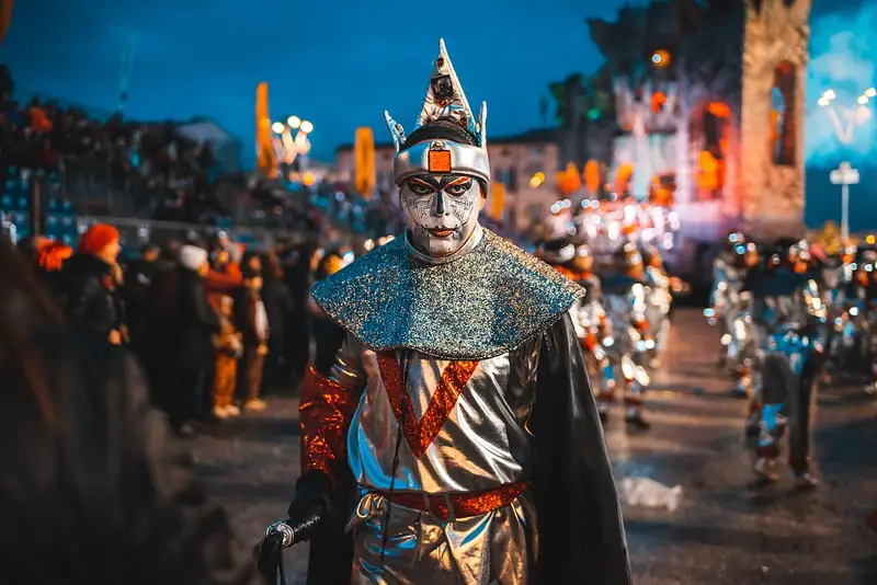 De nuit, un masque costumé en Pierrot argenté défile devant la foule et des chars illuminés