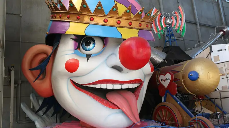 Dans un hangar, une énorme tête de clown en papier mâché peint, couronnée et tirant la langue, attend d'être montée sur un char